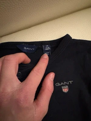 Mörkblått bodyset från GANT - Snyggt mörkblått set från GANT med långärmad body och matchande byxor. Båda plaggen har GANT-logga och emblem broderat på bröstet och höften. Bodyn har praktiska tryckknappar på sidan för enkel påklädning. Perfekt för en stilren och bekväm look.