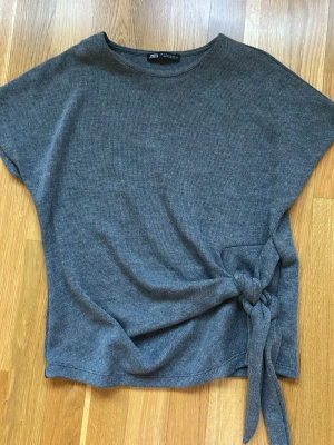 Grå topp med knyt från Zara - Säljer en grå topp från Zara med korta ärmar och rund halsringning. Toppen har en snygg knyt-detalj i sidan som ger en cool twist till outfiten. Perfekt att matcha med både jeans och kjol för en chill look.