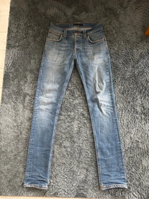 Ljusblå Grim Tim jeans från Nudie - Säljer ett par ljusblå Grimtim slim jeans från Nudie med klassisk femficksdesign och slitna detaljer. Jeansen har oranga sömmar och Nudie-logga på bakfickan. Tillverkade i mjuk denim som sitter tajt hela vägen ner.