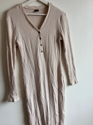 Beige ribbad långklänning med knappar - Långärmad ribbad klänning i beige med v-ringning och knappar framtill. Klänningen har en kroppsnära passform. Perfekt för en stilren och enkel look.