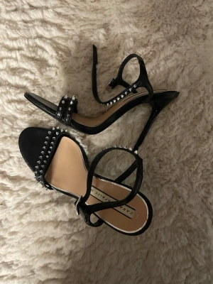Svarta pumps med nitar från Zara - Snygga svarta pumps från Zara med smala remmar och silverfärgade nitar längs kanterna. Skorna har öppen tå och hög klack, samt justerbart spänne runt ankeln. 