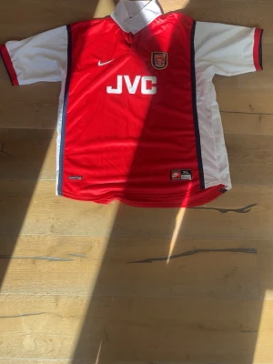 Arsenal retro matchtröja Nike XL - Klassisk Arsenal matchtröja från 1998/1999 med JVC-logga på bröstet och klubbmärke. Tröjan har korta ärmar, vit krage och är tillverkad i lätt och ventilerande polyester. Perfekt för fotbollsträning eller som samlarobjekt.