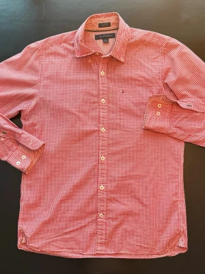 Tommy Hilfiger skjorta - Snygg röd- och vitrutig skjorta från Tommy Hilfiger med klassisk krage och långa ärmar. Skjortan har knappar framtill och en liten logga på bröstfickan. Tillverkad i mjuk bomull som känns skön mot huden.