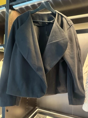 Svart kappa med bred krage - Stilren svart kappa med oversized, bred sjalkrage och rak passform. Kappan är midjelång och har långa ärmar. Tillverkad i ett mjukt, tjockare tyg som ger en elegant look. Perfekt för dig som gillar minimalistisk och modern stil.