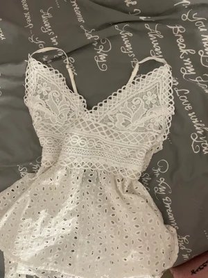 Vit spetslinne med broderi - Säljer ett vitt linne med tunna axelband och vackra spetsdetaljer. Toppen har broderade blommor, hålmönster och volang nedtill. V-ringad fram och knytning i ryggen för en söt och somrig look. Perfekt till jeans eller kjol!