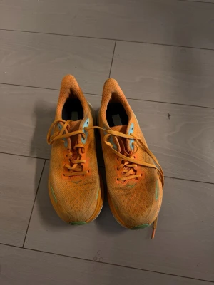 Orange löparskor från Hoka One One - Säljer ett par snygga orange löparskor från Hoka One One. Skorna har mesh-ovandel för bra ventilation, turkosa detaljer och klassisk snörning. Perfekta för dig som vill ha lätta och bekväma skor till löpning eller träning.