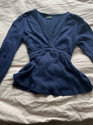 Mörkblå omlottopp med knytband - Snygg mörkblå omlottopp med långa ärmar och v-ringning. Toppen har ett brett knytband i midjan som ger en markerad siluett och ett mjukt, lätt texturerat tyg. Perfekt att styla med jeans eller kjol för en trendig look. Använt 3 gånger och köpte den för 299 kr men säljer den för 130 kr 