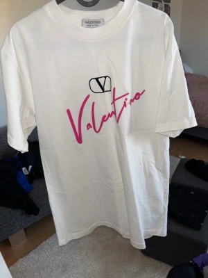 Vit Valentino t-shirt med rosa tryck - Vit t-shirt från Valentino med ett stort rosa logotryck och svart emblem på bröstet. Klassisk passform med rund hals och korta ärmar. Tillverkad i mjuk bomull som känns skön mot huden. Perfekt för dig som gillar stilrena plagg med en twist.