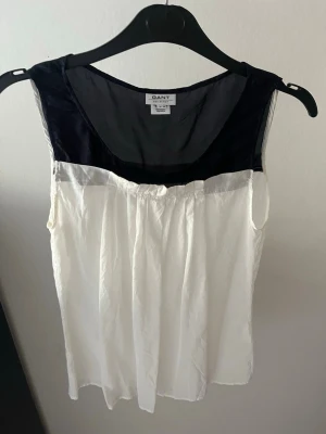 Svartvit linnetopp från Gant - Snygg svartvit linnetopp från Gant med transparent svart axelparti och rund halsringning. Toppen är ärmlös och har en lös passform, perfekt för varma dagar. Materialet känns lätt och luftigt, vilket gör den superbekväm att bära.