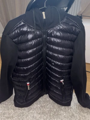 Svart cardiganjacka  - Snygg svart cardiganjacka med quiltad, glansig front och mjuka ärmar. Jackan har huva, dragkedja framtill och två fickor med dragkedja och detaljer. Perfekt för lager på lager.