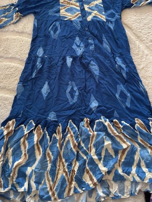 Blå mönstrad långklänning från MMR - Säljer en långklänning från MMR i blått med beige och vita detaljer. Klänningen har ett unikt mönster, knappar framtill och långa ärmar. Perfekt för dig som gillar bohemisk stil och vill sticka ut med färg och form.