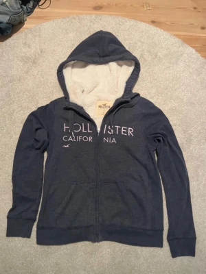 Blå Hollister hoodie med foder - Mysig blå hoodie från Hollister med dragkedja och vit fluffig foder på insidan. Stor logga och text 'Hollister California' på bröstet. Två fickor framtill och klassisk huva. Perfekt för kalla dagar och chill outfits.
