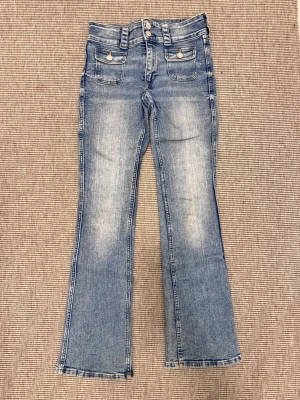 Blå bootcut jeans med dubbla knappar - Snygga ljusblå bootcut jeans med dubbla knappar i midjan och två framfickor med lock. Jeansen har en lätt tvättad look och utsvängda ben. Perfekta för en avslappnad och trendig stil.