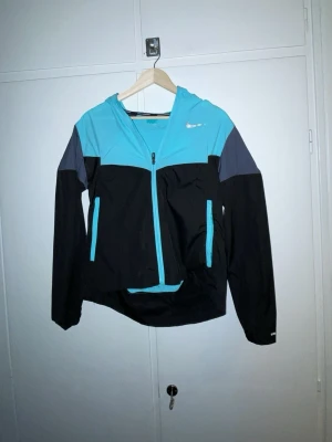 Nike blå och svart vindjacka med huva - Snygg vindjacka från Nike i blått, svart och grått. Jackan har dragkedja framtill, två fickor med dragkedja och en praktisk huva. Perfekt för blåsiga dagar och sportiga outfits. Lätt material som andas och ger en sportig look.