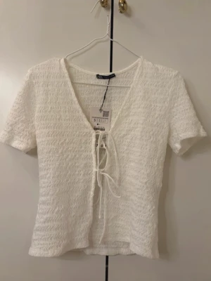 Vit knyt topp Zara - Söt vit topp från Zara med ribbad struktur och korta ärmar. Toppen har v-ringning och knyt framtill som ger en trendig look. Perfekt till jeans eller kjol för en fräsch stil.