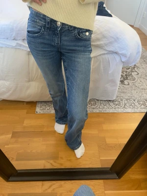 Blå bootcut jeans med paljett-detalj - Säljer ett par blå bootcut jeans med snygg paljett-detalj i form av en rosett på bakfickan. Jeansen har klassisk femficksmodell, silverfärgade knappar och är tillverkade i denim. Perfekta för dig som vill ha en cool och trendig look.