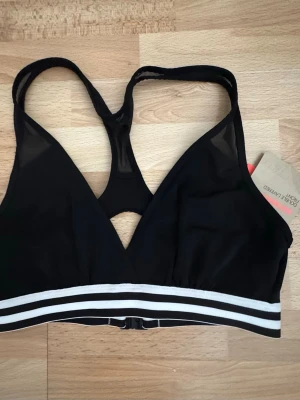 Ny. Svart sport-bh med mesh och ränder - Sportig/swimming svart bh med meshdetaljer och bred resår med vita ränder nedtill. Bh:n har brottarrygg och stängning bak. Materialet är stretchigt och bekvämt, perfekt för en aktiv livsstil.