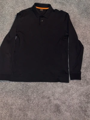 Svart långärmad pikétröja från BOSS - Snygg svart långärmad pikétröja från BOSS med klassisk krage och två knappar framtill. Diskret BOSS-logga broderad på bröstet och orange detalj på insidan av kragen. Perfekt för en stilren look.