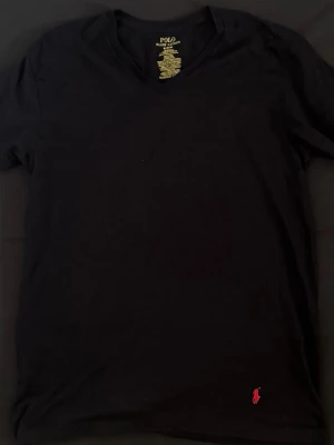 Svart t-shirt från Polo Ralph Lauren - Klassisk svart t-shirt från Polo Ralph Lauren med v-ringning och den ikoniska röda logotypen broderad nertill. Tillverkad i mjuk bomull och har korta ärmar. Perfekt basplagg med stilren design.