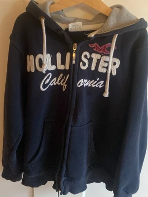 Marinblå hoodie från Hollister - Mysig marinblå hoodie från Hollister med dragkedja,  perfekt för dig som gillar archive stilen.stor broderad logga och text på bröstet. Grå insida på huvan och vita snören. Två fickor framtill och ribbade muddar. Perfekt för en avslappnad och cool stil.