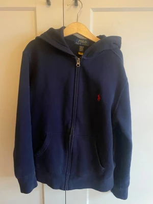 Mörkblå hoodie från Polo Ralph Lauren - Mörkblå hoodie med dragkedja från Polo Ralph Lauren. Klassisk design med huva, kängurufickor och det ikoniska röda Polo-märket broderat på bröstet. Tillverkad i mjuk bomullsblandning som är skön att ha på sig. Perfekt för en avslappnad och stilren look.