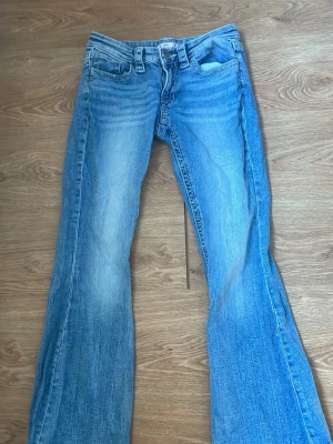 Blå bootcut jeans från Gina Tricot - Säljer ett par klassiska blå bootcut jeans från Gina Tricot med få slitningar vid botten av benen. Jeansen har tydliga sömmar, fyra fickor och ikoniska bakfickor med broderade detaljer. Perfekta för en avslappnad och trendig look.