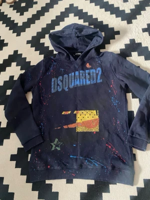 Mörkblå DSQUARED2 hoodie med tryck - Snygg mörkblå hoodie från DSQUARED2 med färgstänk och grafiska tryck i blått, gult, grönt och rött. Stor logga på bröstet och coola detaljer framtill. Hoodien har huva och ribbade muddar vid ärmar och nederkant. Perfekt för dig som gillar streetwear och unika plagg.