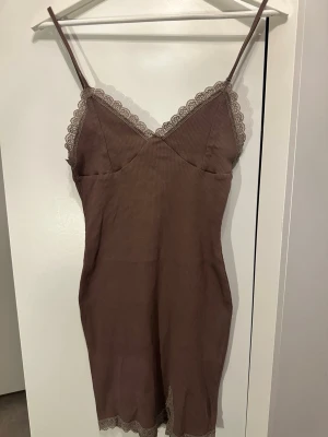 Brunt nattlinne med spetsdetaljer - Snyggt brunt nattlinne med tunna axelband och spetsdetaljer vid urringning och nederkant. Figurnära modell i mjukt, stretchigt tyg som känns skönt mot huden. Perfekt för mysiga kvällar hemma.