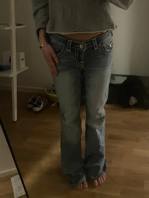 True Religion bootcut jeans ljusblå - Säljer ett par ljusblå bootcut jeans från True Religion med kontrastsömmar och detaljerade fickor. Jeansen har låg midja och klassisk femficksdesign. Perfekta för dig som gillar en avslappnad men ändå trendig stil.