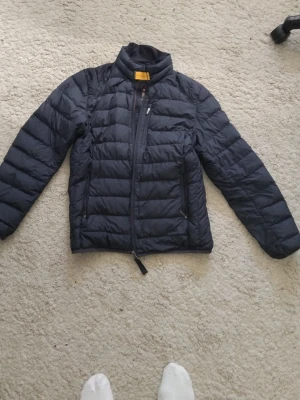 Mörkblå pufferjacka från Parajumpers - Snygg mörkblå pufferjacka från Parajumpers med hög krage och dragkedja framtill. Jackan har två sidofickor med dragkedja och en bröstficka. Tillverkad i ett lätt och värmande material, perfekt för kalla vinterdagar.