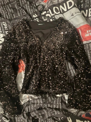 Svart paljettopp från Gina  - Säljer en svart topp från Gina täckt med glittriga paljetter. Toppen har v-ringning och långa ärmar, perfekt för att sticka ut. Materialet känns mjukt och följsamt mot huden. Snygg att matcha med både jeans och kjol.