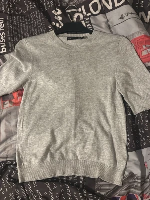 Ljusgrå stickad tröja H&M - En enkel och stilren ljusgrå stickad tröja från H&M. Tröjan har rund halsringning, korta ärmar och ribbade muddar vid ärmslut och nederkant. Perfekt basplagg till garderoben.