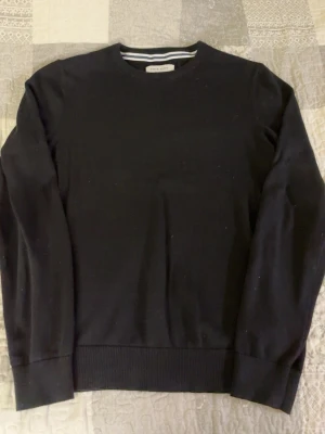 Svart stickad tröja från Pier One - En klassisk svart stickad tröja från Pier One med rund halsringning och ribbade muddar. Perfekt att matcha med jeans eller chinos för en stilren look. Materialet är mjukt och bekvämt, och tröjan har långa ärmar.