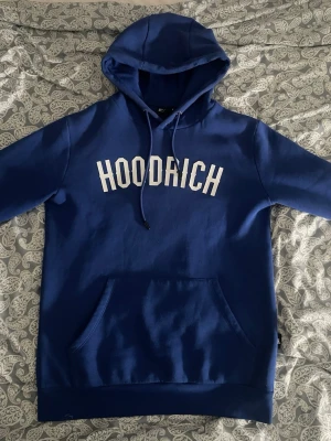 Blå HOODRICH hoodie med tryck - Snygg blå hoodie från Hoodrich med stor vit logga på bröstet och ett coolt tryck på ärmen. Klassisk känguruficka framtill, dragsko i huvan och mjukt material som känns skönt mot huden. Perfekt för dig som gillar streetwear och vill sticka ut.
