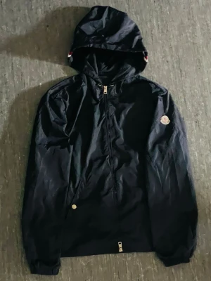 Svart Moncler vindjacka med huva - Säljer en svart vindjacka från Moncler med huva och dragkedja framtill. Jackan har Moncler-logga på vänster ärm och diskreta detaljer i vitt och rött på huvan. Materialet är lätt och vattenavvisande, perfekt för blåsiga dagar.