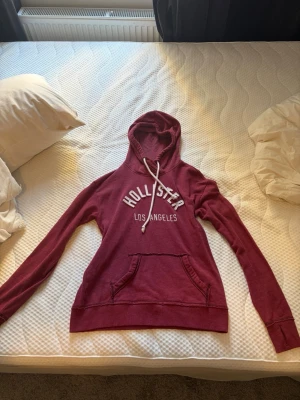 Vinröd hoodie från Hollister - Säljer en vinröd hoodie från Hollister med vit text 'HOLLISTER LOS ANGELES' på bröstet. Hoodien har huva med vita snören och en stor magficka framtill. Långa ärmar med muddar och mjukt material, perfekt för chill dagar.