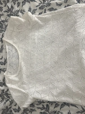 Vit spetsmönstrad topp - Säljer en vit topp med korta ärmar och ett snyggt spetsliknande mönster. Toppen har rund halsringning och är tillverkad i ett mjukt material som känns skönt mot huden. Perfekt att styla med jeans eller kjol för en fräsch look.
