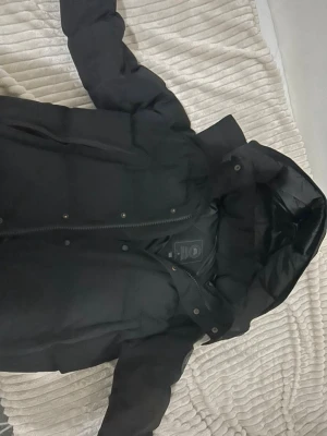 Svart dunjacka från Canada Goose - Säljer en svart dunjacka från Canada Goose med klassisk patch på ärmen. Jackan har huva, dragkedja och tryckknappar framtill. Perfekt för kalla vinterdagar och riktigt varm tack vare dunfyllningen. Snygg och tidlös design som passar till det mesta.