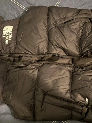 Svart dunväst från The North Face - Snygg svart dunväst från The North Face med dragkedja framtill och två praktiska fickor. Västen har en hög krage och tydlig logga på bröstet och ryggen. Perfekt för lager-på-lager och håller dig varm under kyliga dagar. Storlek L (junior) passar personer som är 160-165 cm långa. Mycket bra skick, säljer för att den inte passar mig längre 