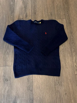 Mörkblå kabelstickad tröja Polo Ralph Lauren - Mörkblå kabelstickad tröja från Polo Ralph Lauren med rund halsringning och klassisk röd logga broderad på bröstet. Tröjan har långa ärmar och ribbade muddar vid ärmslut och nederkant. Perfekt för en stilren och avslappnad look. Storlek M men krympt till XS