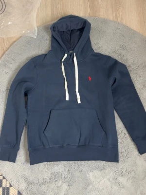 Marinblå hoodie Polo Ralph Lauren - Snygg marinblå hoodie från Polo Ralph Lauren i slim fit-modell. Klassisk känguruficka framtill, vit dragsko i huvan och den ikoniska röda loggan broderad på bröstet. Tillverkad i mjuk bomullsblandning för skön känsla.