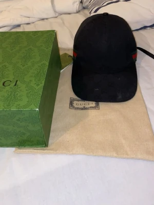 Svart keps från Gucci med logga - Säljer en svart keps från Gucci med diskret GG-monogrammönster och klassiska gröna och röda ränder på sidan. Justerbar rem baktill och böjd skärm. Kepsen kommer med originalförpackning och dustbag. Priset kan diskutera vidare snabba affär 😄