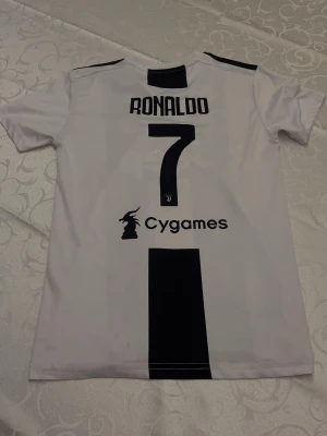 Juventus Ronaldo matchtröja - Juventus hemmatröja från Adidas med Ronaldo och nummer 7 på ryggen. Tröjan är vit med svarta ränder, har Jeep och Cygames som sponsorer samt Juventus klubbmärke och tre stjärnor på bröstet. Tillverkad i lätt och ventilerande material, perfekt för fotbollsträning.