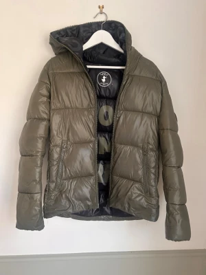 Olivgrön pufferjacka från save the Duxk - Snygg olivgrön pufferjacka med huva och dragkedja framtill. Jackan har två sidofickor med dragkedja och quiltad design. Perfekt för kalla dagar och ger en trendig look. Fodrad insida. Materialet är glansigt och vattenavvisande.