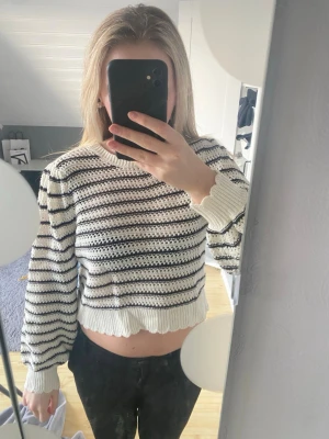 Randig stickad tröja med öppen rygg - Trendig stickad tröja i vitt med svarta horisontella ränder. Croppad modell med långa ärmar och ribbade muddar. Baksidan har öppen design med två knytband upptill och nedtill för en unik look. 