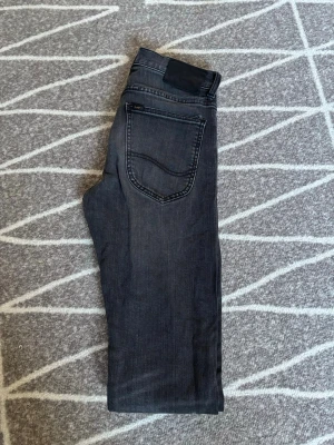 Mörkgrå jeans från Lee - Snygga mörkgrå jeans från Lee med klassisk femficksdesign och svarta detaljer. Jeansen har raka ben och är tillverkade i mjukt denimtyg som ger en skön passform. Perfekta för dig som gillar en stilren och enkel look. Storlek W27 L32. Hör av er vid frågor eller lägg ett prisförslag!