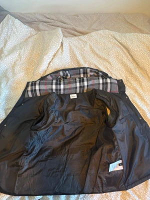 Svart dunväst med rutigt foder - Snygg svart dunväst med dragkedja och tryckknappar framtill. Västen har två fickor med dragkedja och ett klassiskt rutigt foder i grått, vitt och rött. Perfekt för lager-på-lager och ger en stilren look med extra värme.