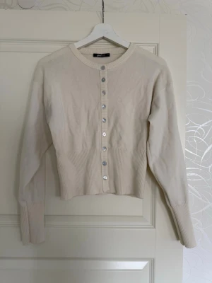 Krämvit kofta från Gina Tricot - Snygg krämvit kofta från Gina Tricot med rund halsringning och silvriga knappar framtill.  Den är i storlek M men passar mer till S.