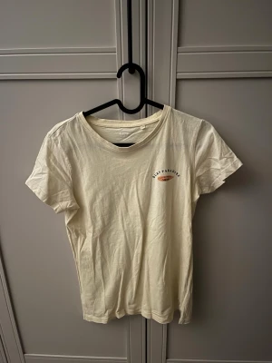 Vit t-shirt med Surf Paradise-tryck - Snygg vit t-shirt med trycket 'Surf Paradise' och palmer på ryggen samt ett mindre tryck fram. Skön passform och klassisk rund hals. Perfekt för dig som gillar avslappnad och cool stil. Tshirten är från lager157 och är i storlek 150