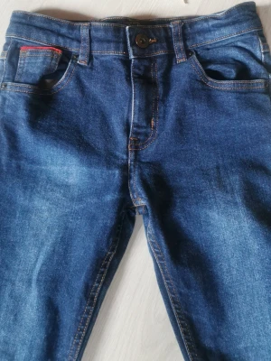 Mörkblå jeans med gul detalj - Snygga mörkblå jeans med klassisk femficksmodell och kontrastsömmar. Jeansen har en liten gul brodyr på ena bakfickan och en röd detalj vid myntfickan fram. Materialet är denim i bomull med lätt tvättad look och raka ben.